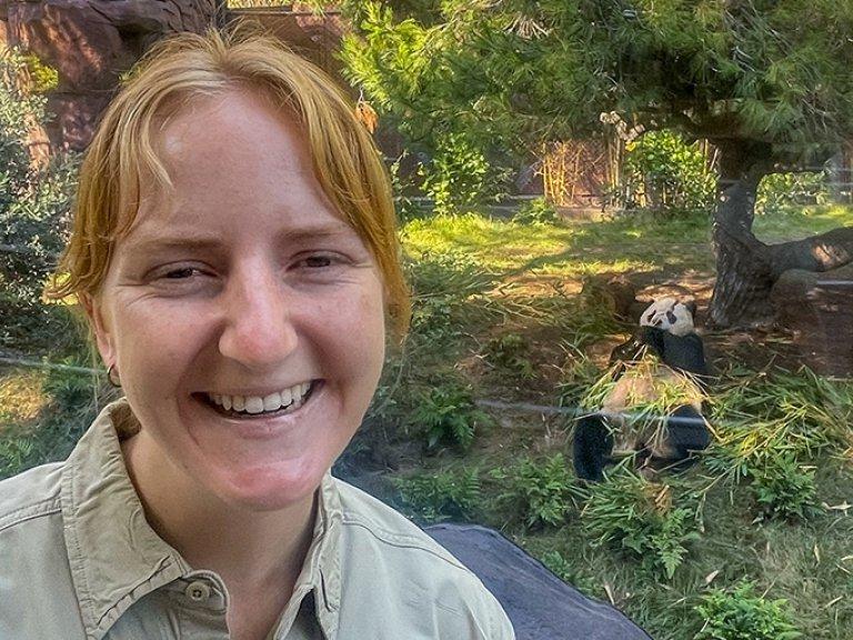 Jana Biedenweg stands in front of the giant panda enclosure at San Diego Zoo while a panda eats bamboo.